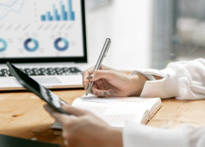 Businesswoman Writing Note And Using Tablet At Her Desk Office. - Barão Assessoria Contábil